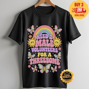 May include: A black short-sleeve t-shirt featuring a colorful graphic print with a rainbow, smiling suns, and butterflies. The text reads NEED 2 MALE VOLUNTEERS FOR A THREESOME in a retro, bubble-style font with pink, purple, and yellow colors.