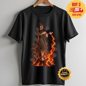 Peut inclure: Un t-shirt noir à manches courtes avec une impression graphique représentant une personne aux longs cheveux sombres attachée à un poteau en bois, entourée de flammes orange et jaunes vives. La personne porte une robe marron et semble cracher du feu.