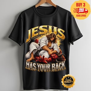 Könnte beinhalten: Ein schwarzes Kurzarm-T-Shirt mit einer Grafik einer Figur in weißem Gewand mit Kreuzhalskette, die mit einer roten gehörnten Kreatur ringt. Der Text JESUS HAS YOUR BACK ist in großen goldenen Buchstaben über und unter dem Motiv gedruckt.