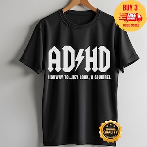 Könnte beinhalten: Ein schwarzes Kurzarm-T-Shirt mit weißem Aufdruck AD/HD, bei dem ein Blitz den Schrägstrich ersetzt. Unter dem Haupttext zeigt das T-Shirt den Schriftzug HIGHWAY TO...HEY LOOK, A SQUIRREL in einer klaren, serifenlosen weißen Schriftart.