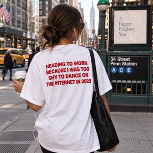 Puede incluir: Una camiseta blanca de manga corta con texto rojo en la parte posterior que dice: HEADING TO WORK BECAUSE I WAS TOO SHY TO DANCE ON THE INTERNET IN 2020. La prenda se lleva con un bolso de hombro negro en un entorno urbano.
