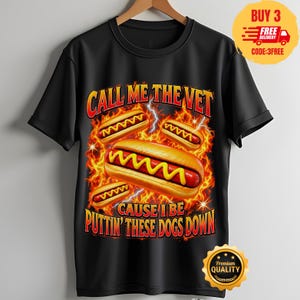 Op de afbeelding: Een zwart t-shirt met korte mouwen met een afbeelding van hotdogs met gele mosterd tegen een achtergrond van oranje en rode vlammen en bliksem. De tekst luidt CALL ME THE VET CAUSE I BE PUTTIN' THESE DOGS DOWN in gele en rode letters.