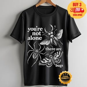 Puede incluir: Una camiseta negra de manga corta con un estampado gráfico en blanco de un escarabajo, una araña y una polilla. El texto en la camiseta dice, you're not alone there are bugs. El diseño está centrado en la parte delantera de la prenda.