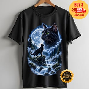 May include: A black short-sleeved t-shirt featuring a graphic print of a large black cat head with green eyes overlooking a smaller black cat standing on a rocky cliff above a waterfall, surrounded by a pine forest and a glowing full moon.