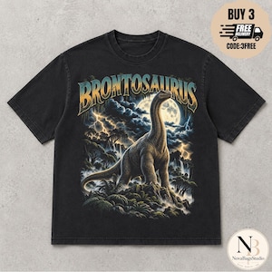 Pode incluir: Uma t-shirt preta de manga curta com uma estampa gráfica de um Brontossauro num cenário pré-histórico com lua cheia, relâmpagos e vegetação exuberante. A palavra BRONTOSAURUS está impressa numa fonte grande e estilizada acima do dinossauro.