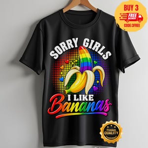 Może przedstawiać: Czarny t-shirt z krótkim rękawem z grafiką częściowo obranego banana z tęczową górą. Napis głosi SORRY GIRLS I LIKE BANANAS. Wzór zawiera kolorowe tło w kropki z małymi sercami i gwiazdkami, umieszczone centralnie na przodzie koszulki.