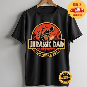 Op de afbeelding: Een zwart t-shirt met korte mouwen en een cirkelvormige afbeelding op een oranje achtergrond. Het ontwerp toont een skelet dat zijn onderrug vasthoudt, een vulkaan en palmbomen. De tekst luidt JURASSIC DAD en PAIN FINDS A WAY in vetgedrukte letters.