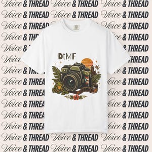 Może przedstawiać: Biały t-shirt z krótkim rękawem z nadrukiem graficznym przedstawiającym aparat fotograficzny, małą żabę, tropikalne liście, zachód słońca oraz napis DtMF. Wzór zawiera element taśmy filmowej i mały detal w postaci flagi na korpusie aparatu.