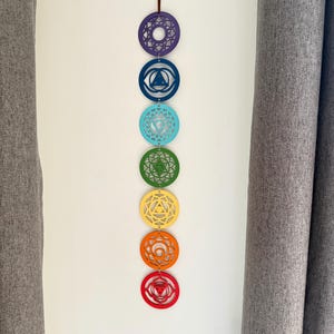 May include: A colorful chakra wall hanging. Seven circular wooden ornaments in a vertical row, each a different color: purple, blue, turquoise, green, yellow, orange, and red. Each circle has a unique geometric design. A brown string is at the top for hanging.
