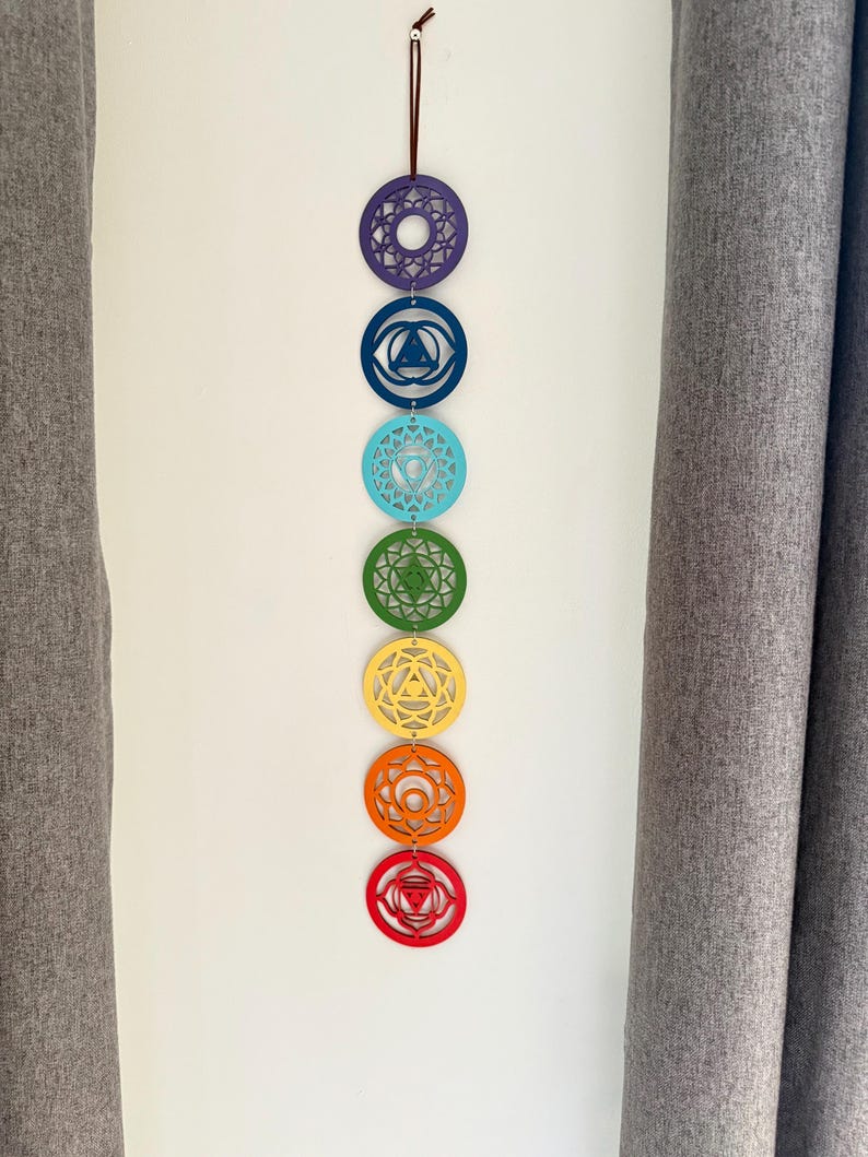 May include: A colorful chakra wall hanging. Seven circular wooden ornaments in a vertical row, each a different color: purple, blue, turquoise, green, yellow, orange, and red. Each circle has a unique geometric design. A brown string is at the top for hanging.