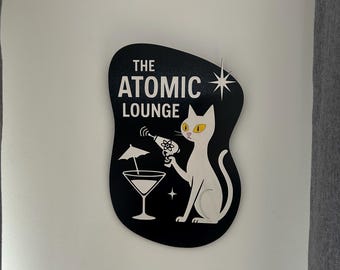 May include: Black and white illustration of a cat holding a martini glass with an umbrella. The cat is wearing a ray gun and has a star above its head. The text "THE ATOMIC LOUNGE" is written in white letters on a black background.