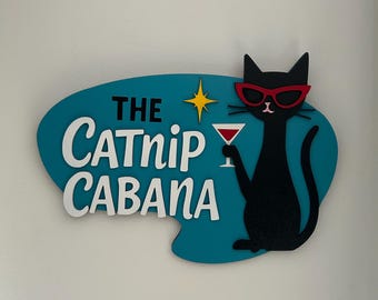 May include: A teal blue sign with a white oval shape and the text "THE CATNIP CABANA" in white lettering. A black cat with red sunglasses is holding a red cocktail glass. A white star is in the top right corner of the sign.