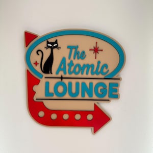 May include: A vintage-style sign for "The Atomic Lounge" with a black cat, a red arrow, and teal and beige colours. The sign has a mid-century modern design, suitable for a bar or lounge setting.