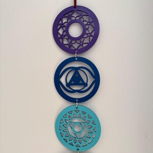 May include: A vertical display of four colorful chakra symbols, each a different shade of blue, green, and purple. The circular designs are suspended by a maroon string, with intricate cut-out patterns within each circle. The background is a plain white wall.