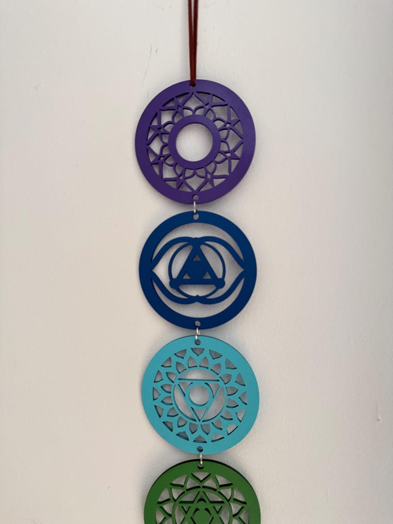 May include: A vertical display of four colorful chakra symbols, each a different shade of blue, green, and purple. The circular designs are suspended by a maroon string, with intricate cut-out patterns within each circle. The background is a plain white wall.