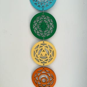 May include: A vertical string of five colorful wooden chakra symbols. The top circle is teal, followed by green, yellow, orange, and red. Each circle has a unique geometric design and is connected by small metal rings.