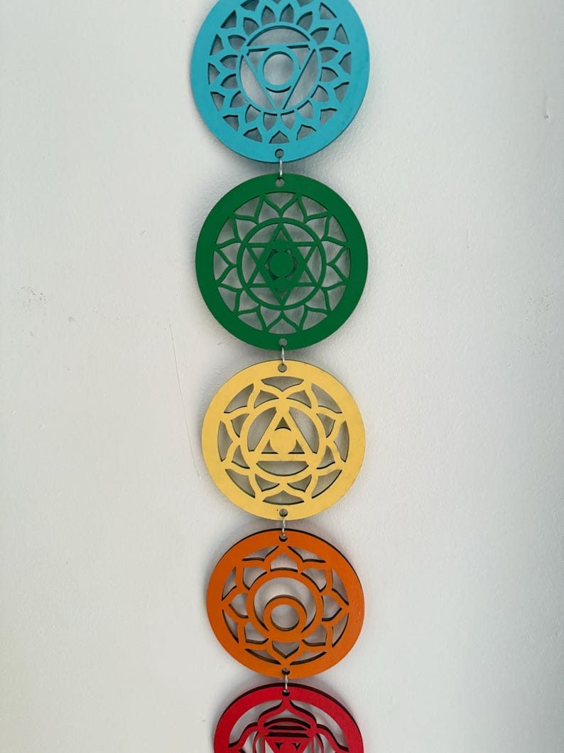 May include: A vertical string of five colorful wooden chakra symbols. The top circle is teal, followed by green, yellow, orange, and red. Each circle has a unique geometric design and is connected by small metal rings.