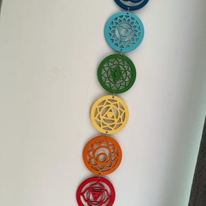 May include: A vertical display of seven colorful chakra symbols. Each symbol is a different color, including red, orange, yellow, green, blue, and purple. The circular symbols are connected by small metal rings.
