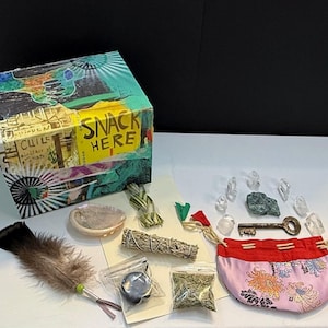 Può includere: Una collezione di oggetti spirituali tra cui una scatola decorata con la scritta "SNACK HERE", cristalli, una piuma, una chiave, un piccolo sacchetto floreale rosa e mazzi di erbe. La scatola è adornata con elementi di collage colorati.