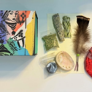 Può includere: Un assortimento di articoli spirituali tra cui una scatola con un disegno di regina di picche, mazzi di salvia, una piuma, cristalli, una conchiglia e un sacchetto rosso con un motivo geometrico.