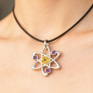 May include: Silver-tone pendant necklace featuring an atom design. The pendant has a yellow center stone and three purple stones. The necklace is on a black cord.