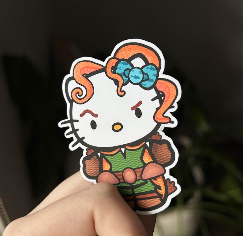 May include: A sticker depicting a cartoon cat character with orange hair and a blue bow. The cat is dressed in a green and brown outfit with orange details. The cat has an angry expression.