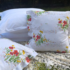 May include: White pillowcases with a floral pattern of red and yellow flowers and green stems. The pillowcases have a ruffled edge. A stack of white pillowcases is in the background.