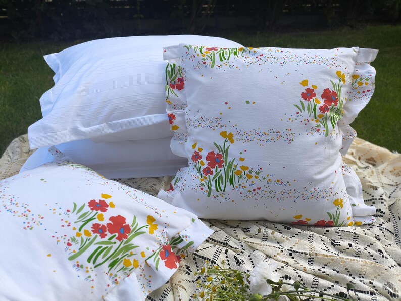 May include: White pillowcases with a floral pattern of red and yellow flowers and green stems. The pillowcases have a ruffled edge. A stack of white pillowcases is in the background.