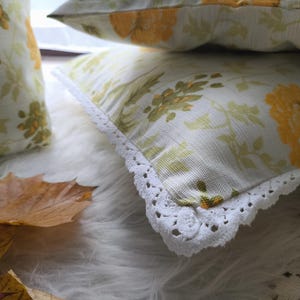 May include: Close-up of decorative pillows with a floral pattern in yellow and green on a white background. The pillows have a white lace trim. A few autumn leaves are in the foreground.