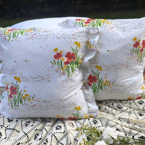 May include: Two white throw pillows with a floral pattern of red and yellow flowers and green stems. The pillows have ruffled edges and are placed on a white lace tablecloth. The pillows are decorated with colorful speckles.
