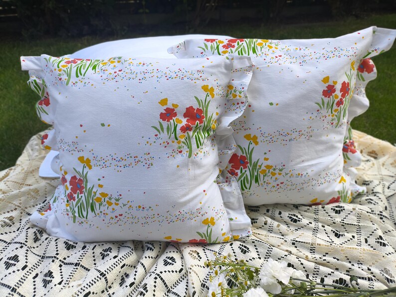May include: Two white throw pillows with a floral pattern of red and yellow flowers and green stems. The pillows have ruffled edges and are placed on a white lace tablecloth. The pillows are decorated with colorful speckles.