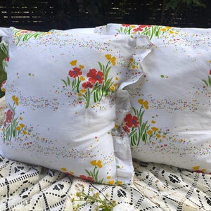 May include: Two white throw pillows with a floral pattern featuring red and yellow flowers, green stems, and colorful speckles. The pillows have ruffled edges and are placed on a white lace tablecloth.