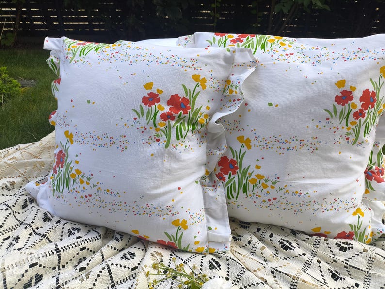 May include: Two white throw pillows with a floral pattern featuring red and yellow flowers, green stems, and colorful speckles. The pillows have ruffled edges and are placed on a white lace tablecloth.
