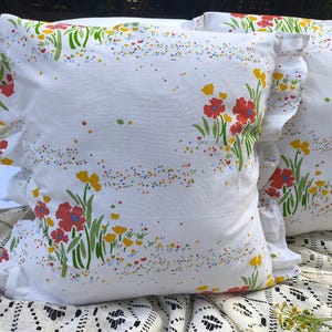 May include: Two white throw pillows with a floral pattern featuring red and yellow flowers, green stems, and colorful speckles. The pillows have ruffled edges and are placed on a white lace cloth.