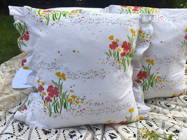 May include: Two white throw pillows with a floral pattern featuring red and yellow flowers, green stems, and colorful speckles. The pillows have ruffled edges and are placed on a white lace cloth.