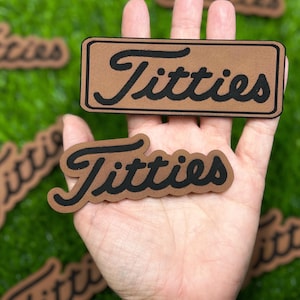 May include: Two brown felt patches with black lettering that reads "Titties" in a cursive font. The patches are held in a hand.