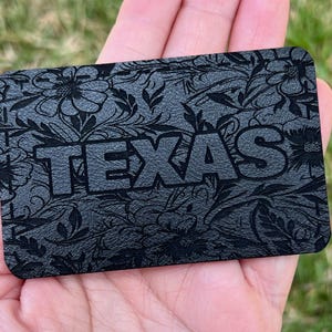 May include: A rectangular, dark gray card with the word "TEXAS" embossed in large, block letters. The card features a detailed floral pattern. The card is held in a person's hand against a blurred green background.