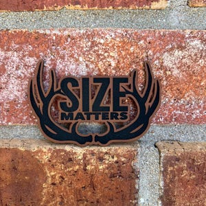 May include: A wooden sign with the words "SIZE MATTERS" in black lettering. The sign is shaped like a pair of antlers with a brown border and is set against a red brick wall.