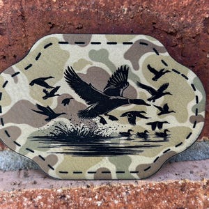 May include: A camouflage-patterned patch with a black silhouette of a flock of ducks flying over a lake. The patch has a stitched border.
