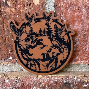 May include: A brown leather patch with a black silhouette of a dog's head and flying ducks. The dog's head is in the center of the patch and the ducks are flying around the dog's head. There are trees in the background.
