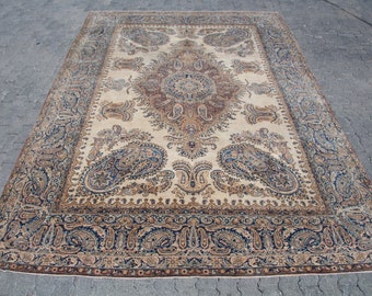 Puede incluir: Una alfombra rectangular con un campo de color crema, un medallón central y complejos patrones de cachemira en tonos azul, marrón y beige. El borde muestra un motivo floral y de cachemira repetitivo en colores a juego.
