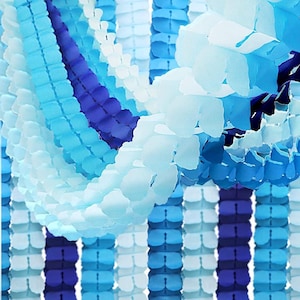 May include: A close-up of blue and white paper decorations. The decorations are in a honeycomb or garland style, with varying shades of blue and white. The paper has a textured appearance, and the overall effect is festive and decorative.