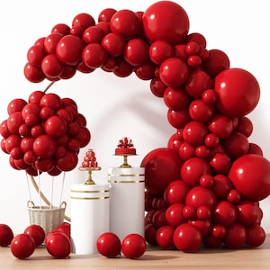 May include: A festive display featuring a red balloon arch, a cluster of red balloons resembling a hot air balloon, and several red balloons scattered on the floor. Two white pedestals hold small red cakes. A wicker basket is also present.