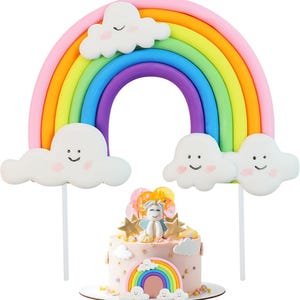 May include: Rainbow cake topper set with a rainbow arch, clouds, and a unicorn. The rainbow arch is made of multiple colors, including pink, orange, yellow, green, blue, and purple. The clouds are white with pink accents and have happy faces. The unicorn is white with gold accents and is sitting on a pink cake.