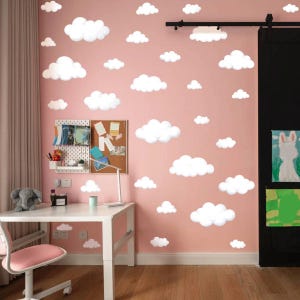 May include: A pink wall decorated with white cloud-shaped decals. The room features a white desk, a pink chair, and a small shelf with various items. A black curtain and artwork are visible on the right side. The room has a cozy and playful atmosphere.