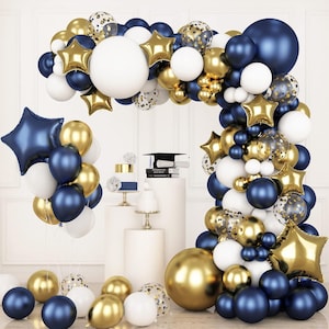 May include: A festive balloon arch in navy blue, gold, white, and clear. The arrangement includes round and star-shaped balloons, with confetti-filled balloons. A graduation cap and small cakes are also visible, suggesting a celebration.