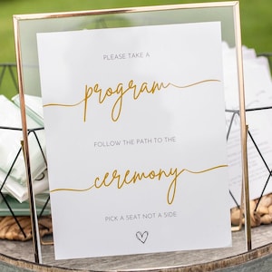 May include: A white sign with gold lettering in a gold frame. The sign says "Please take a program, follow the path to the ceremony, pick a seat not a side".