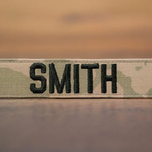 May include: A rectangular patch with the name "SMITH" embroidered in black capital letters. The patch has a camouflage pattern in shades of green, tan, and brown. The patch is likely for military or tactical gear.