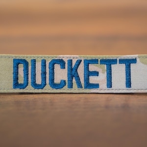 May include: A beige and white camouflage fabric name tag with the name "DUCKETT" embroidered in blue thread.