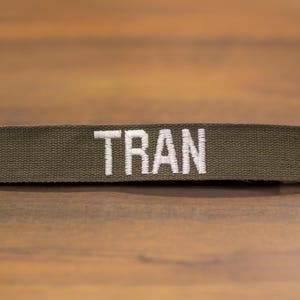 Puede incluir: Cinta de nombre de tela verde oliva con el nombre "TRAN" bordado en letras blancas. La cinta es rectangular y se encuentra horizontalmente sobre una superficie de madera. El nombre está centrado en la cinta.